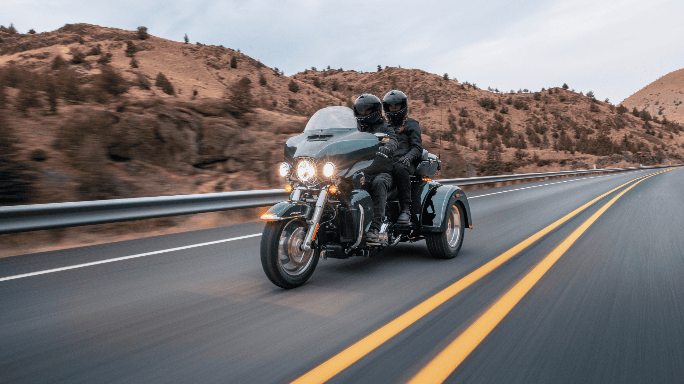 Two riders on a 2024 Harley-Davidson® Tri Glide® Ultra cruising through scenic open roads, showcasin…